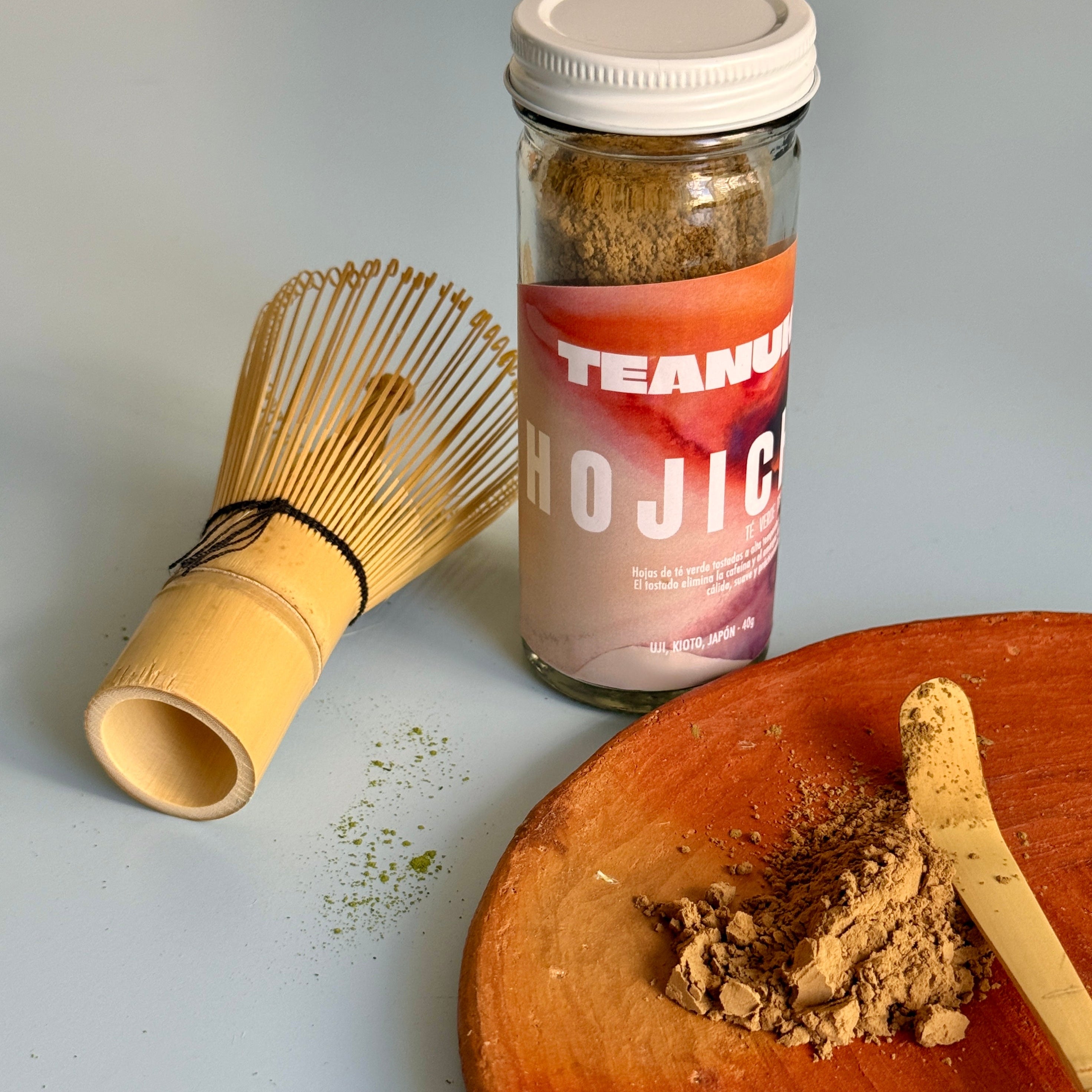 Tea canister labeled 'Hojicha' with a clay plate containing hojicha powder and a whisk on a light blue background.