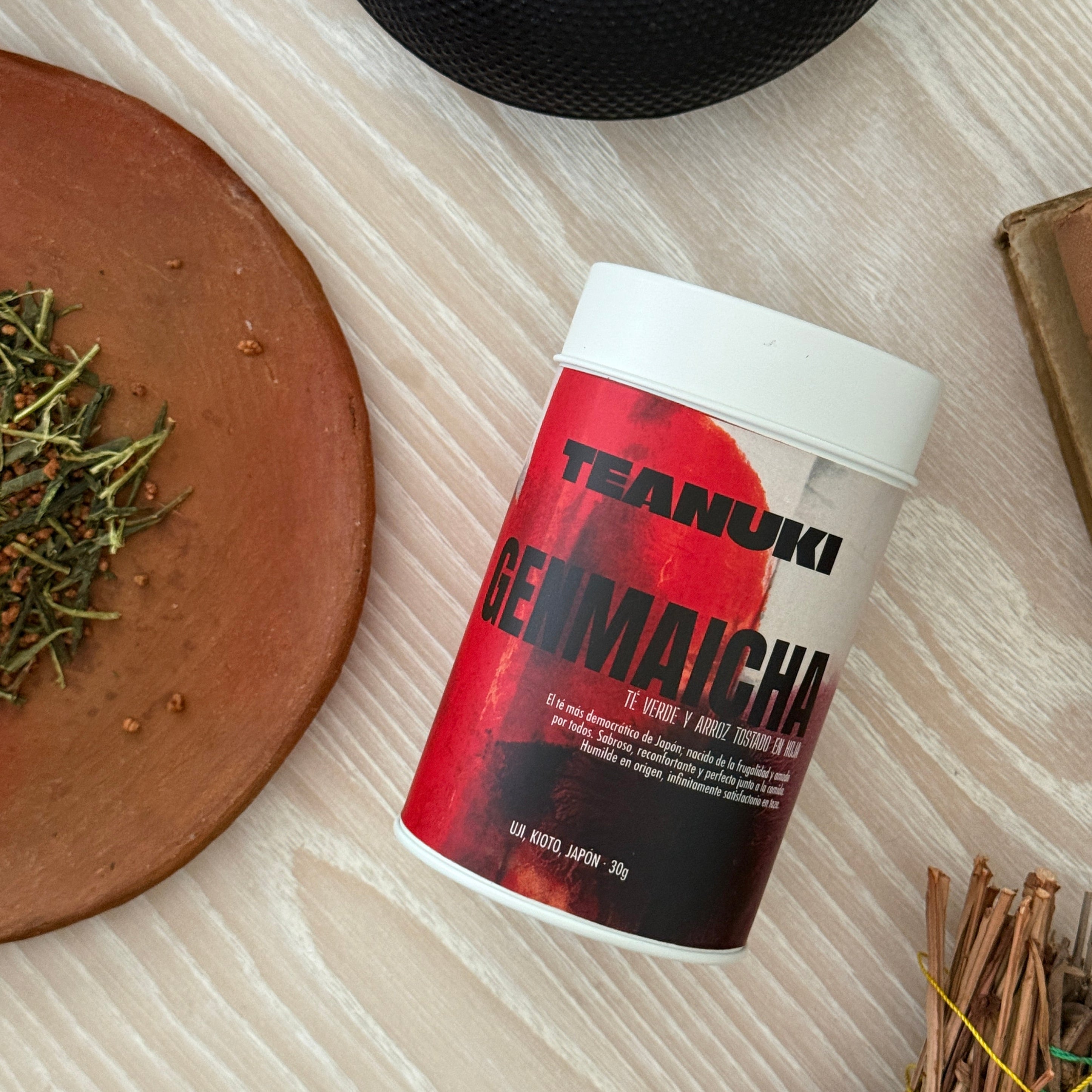 Teanuki genmaicha green tea tin packaging with a red label on a wooden surface with herbs and a black lid.