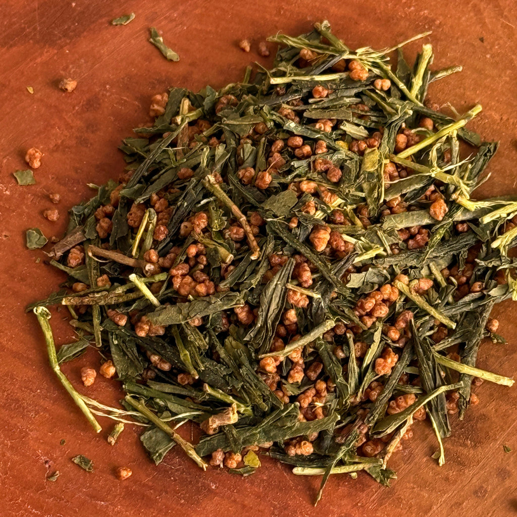 Dry green tea leaves with some brown rice mixed in on a brown clay surface