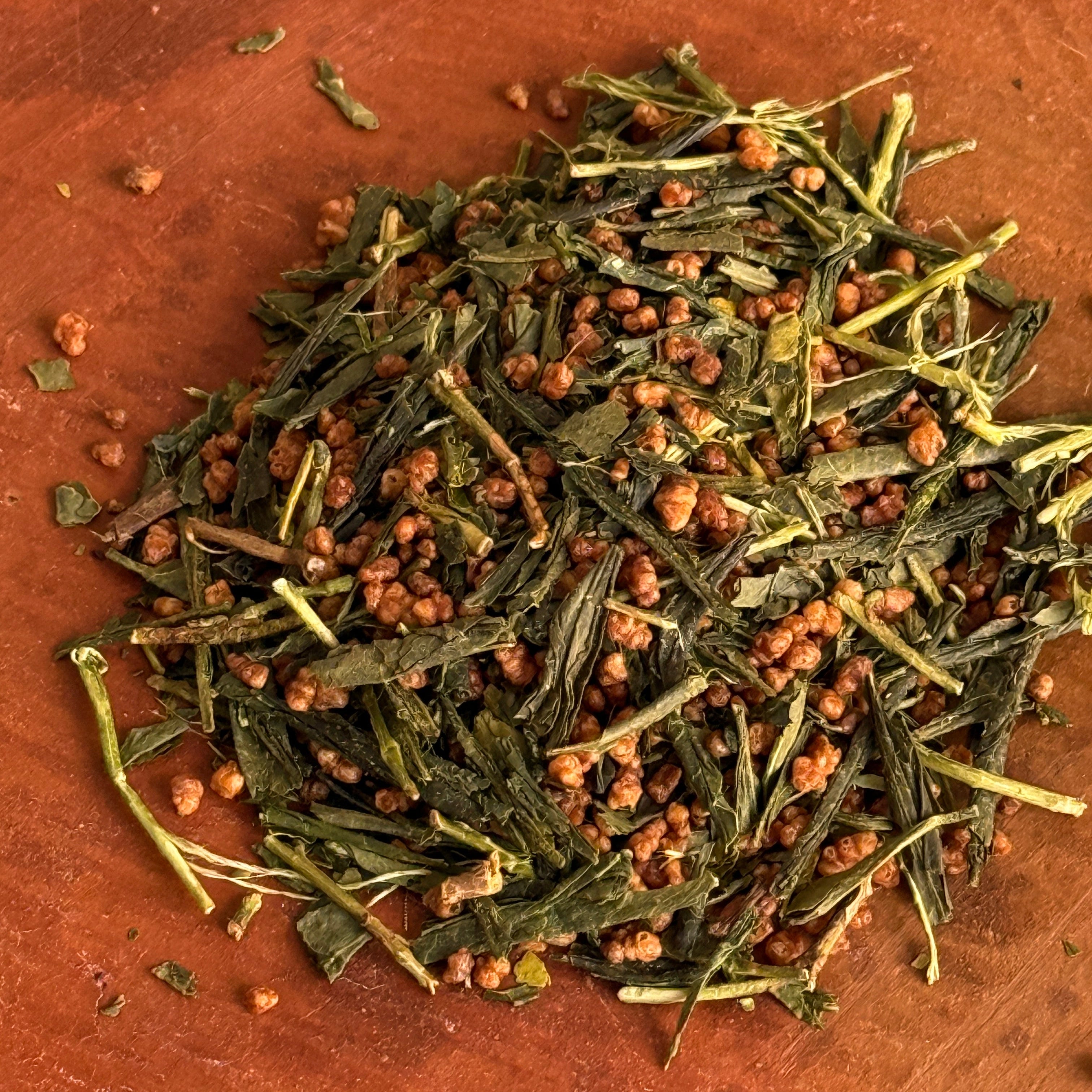 Dry green tea leaves with some brown rice mixed in on a brown clay surface