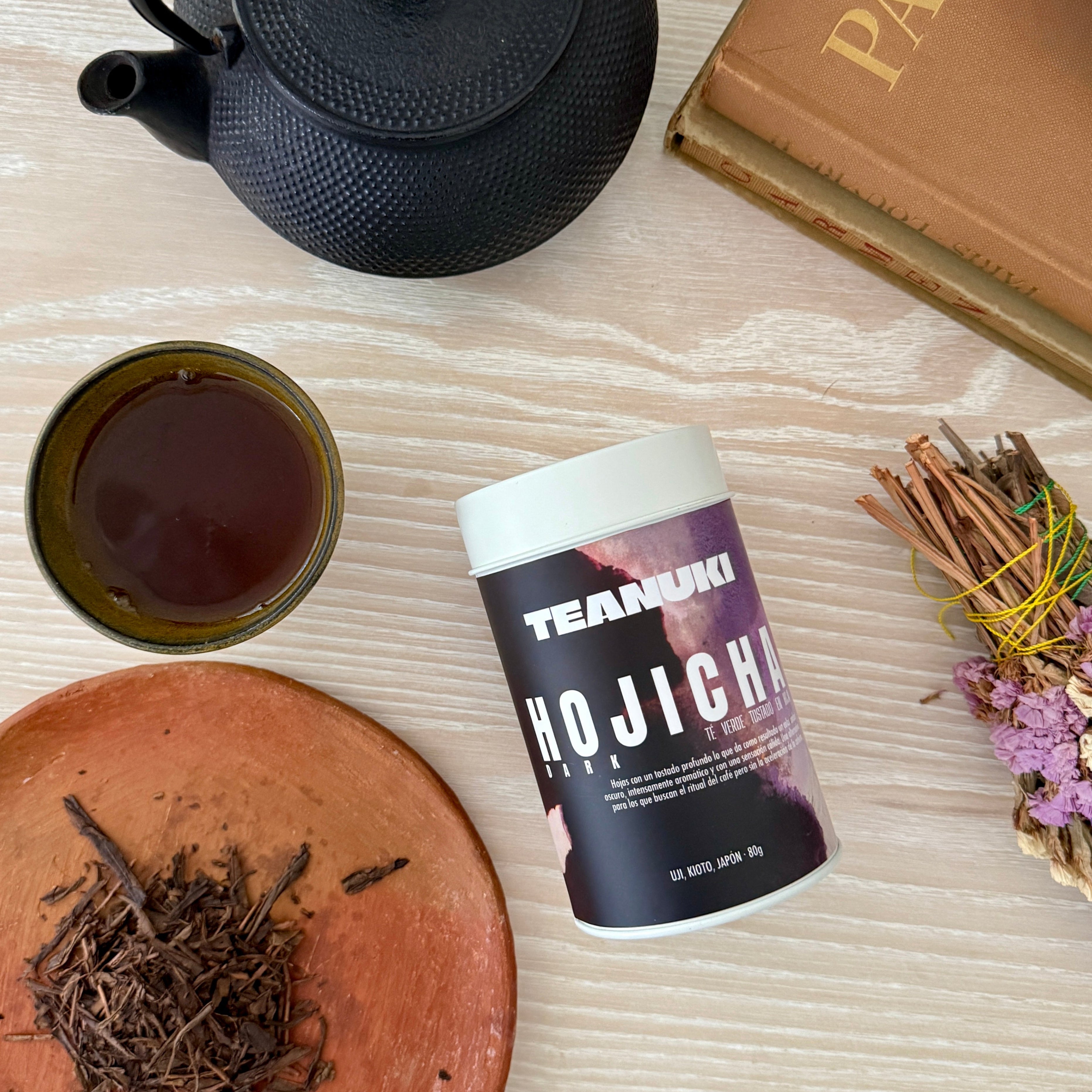 Dark Roasted Japanese Hojicha set with black iron teapot, tea leaves, a canister from Teanuki labeled 'Hojicha', and a book titled 'Paris' on a wooden surface.