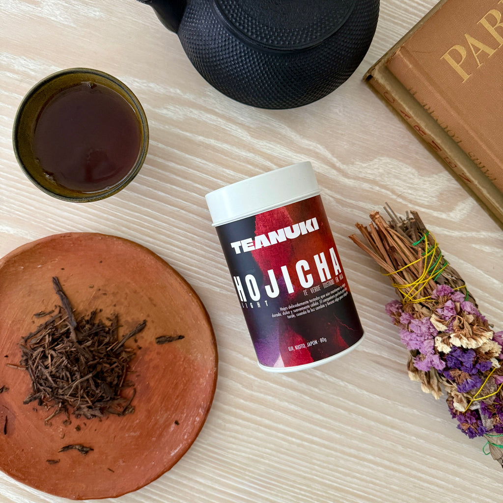 Tea set with a black teapot, cup of tea, TEANUKI Hojicha container, and dried herbs on a wooden surface.