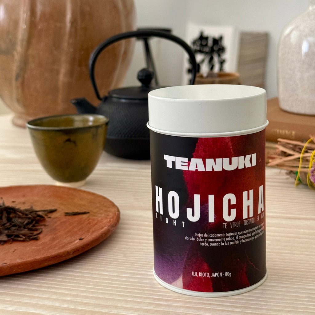 Teanuki Hojicha tea canister on a table with tea leaves and a teapot in the background.